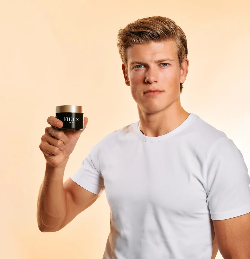 Man in white shirt holding a jar of men's skincare product against a beige background.